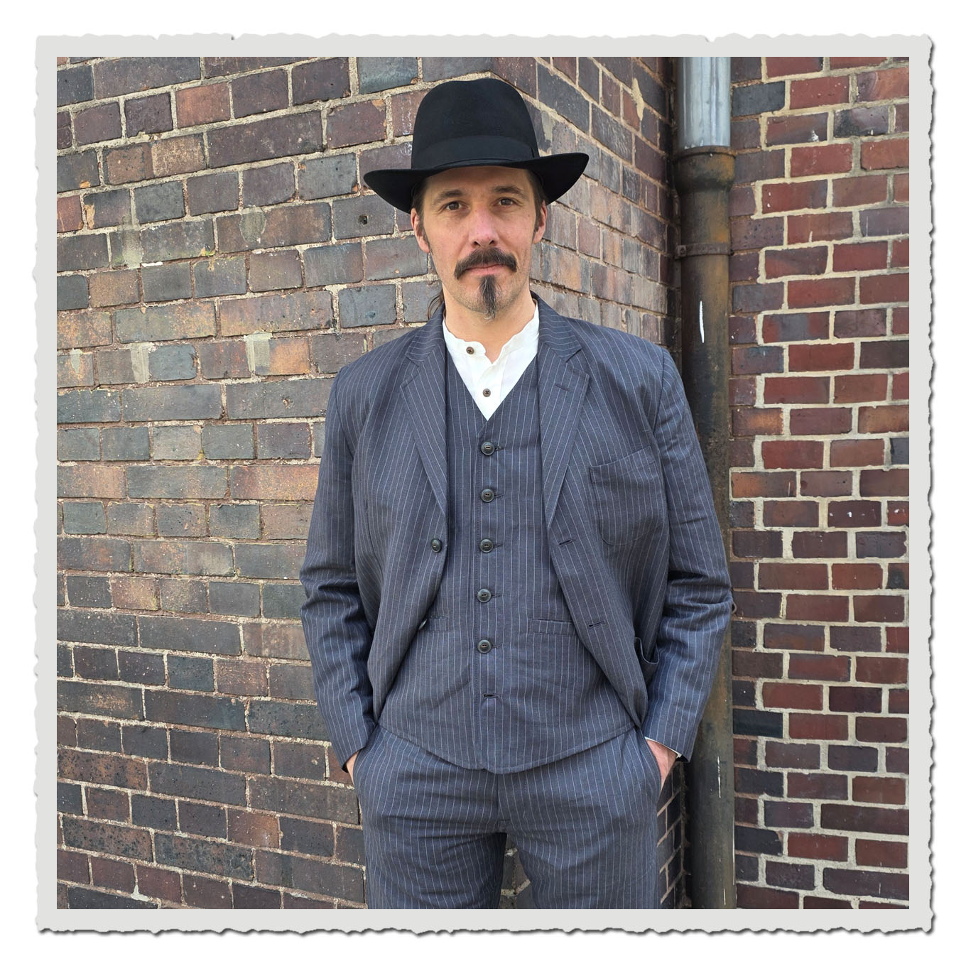 Model in grauem Nadelstreifen-Sakko mit Weste vor dunkler Backsteinwand im Außenbereich.